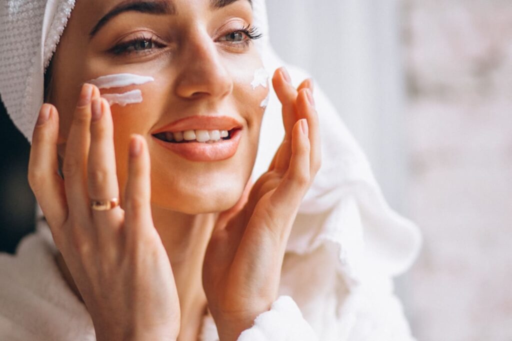 Mulher sorridente aplicando hidratante facial no rosto após o banho, ilustrando a primeira etapa da rotina de skincare recomendada pela dermatologista Dra. Luciane na Clínica Reviva, localizada no Cambuí em Campinas.